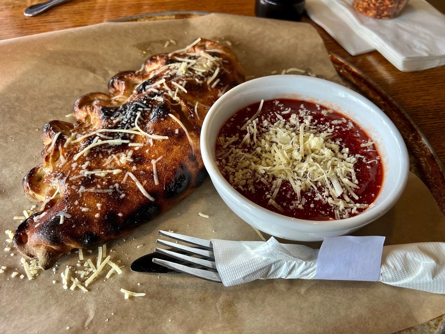 Delicious-looking calzone (in a crescent shape), with a little bit of cheese on top and brown from being baked in butter, next to a little bowl of marinara sauce with cheese in it.