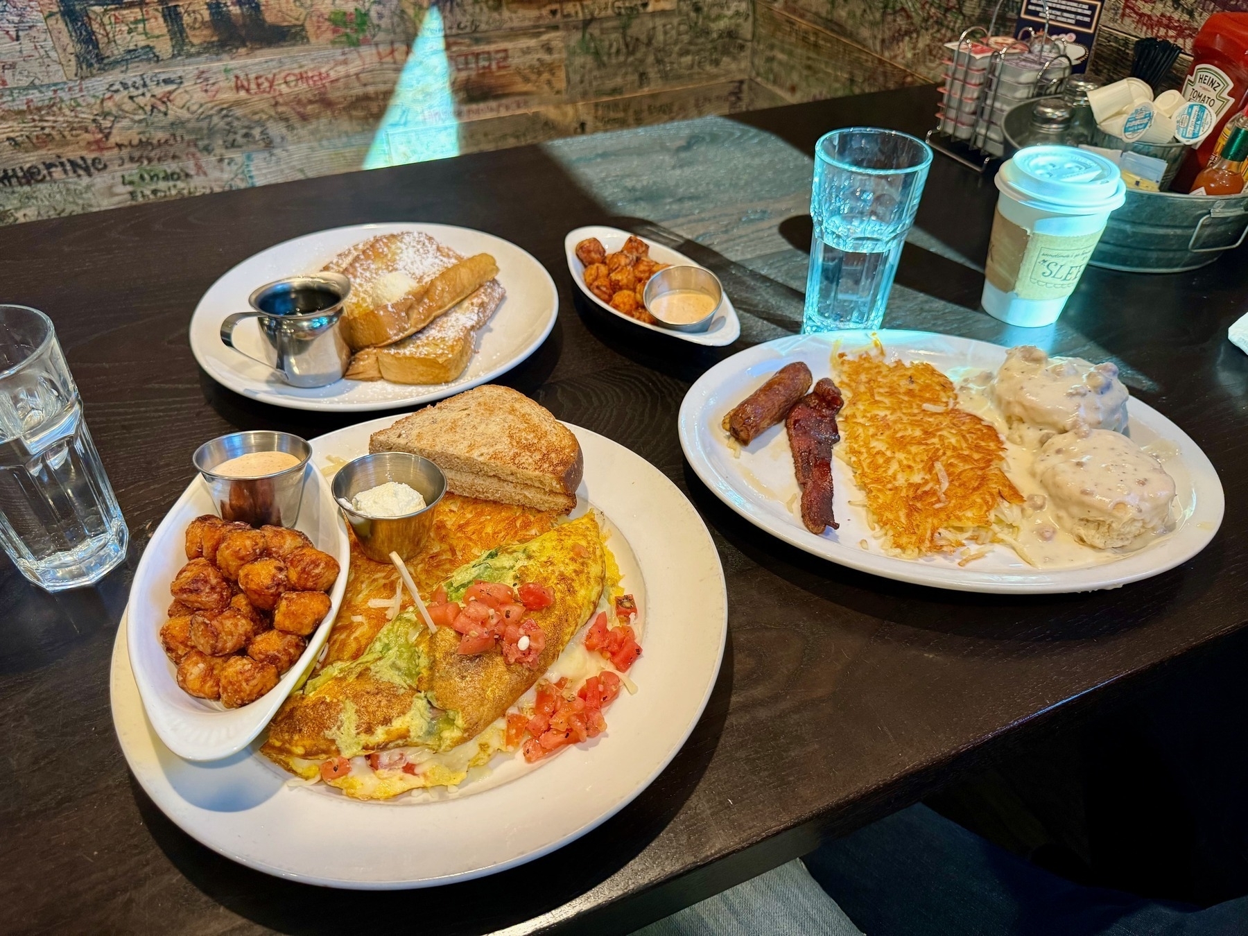 Four plates full of breakfast food, with an omlet, hashbrows, biscuits / gravey, french toast, two servings of tots, and toast...along with two glasses of water and a cup of coffee in a paper cup.