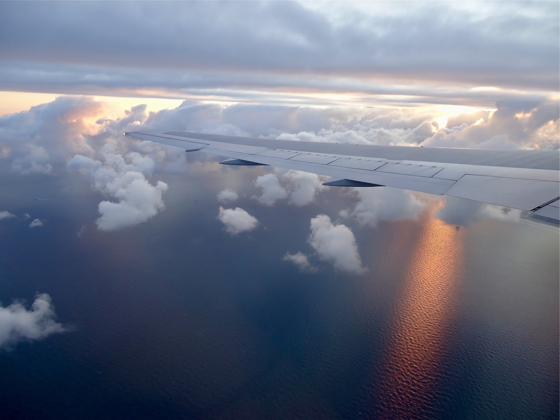iew of the sky and ocean, from an airplane; the ocean is a deep blue/purple below, with a hint of sunlight appearing on the water from behind the clouds, while the white wing of the plan is across the middle of the image.