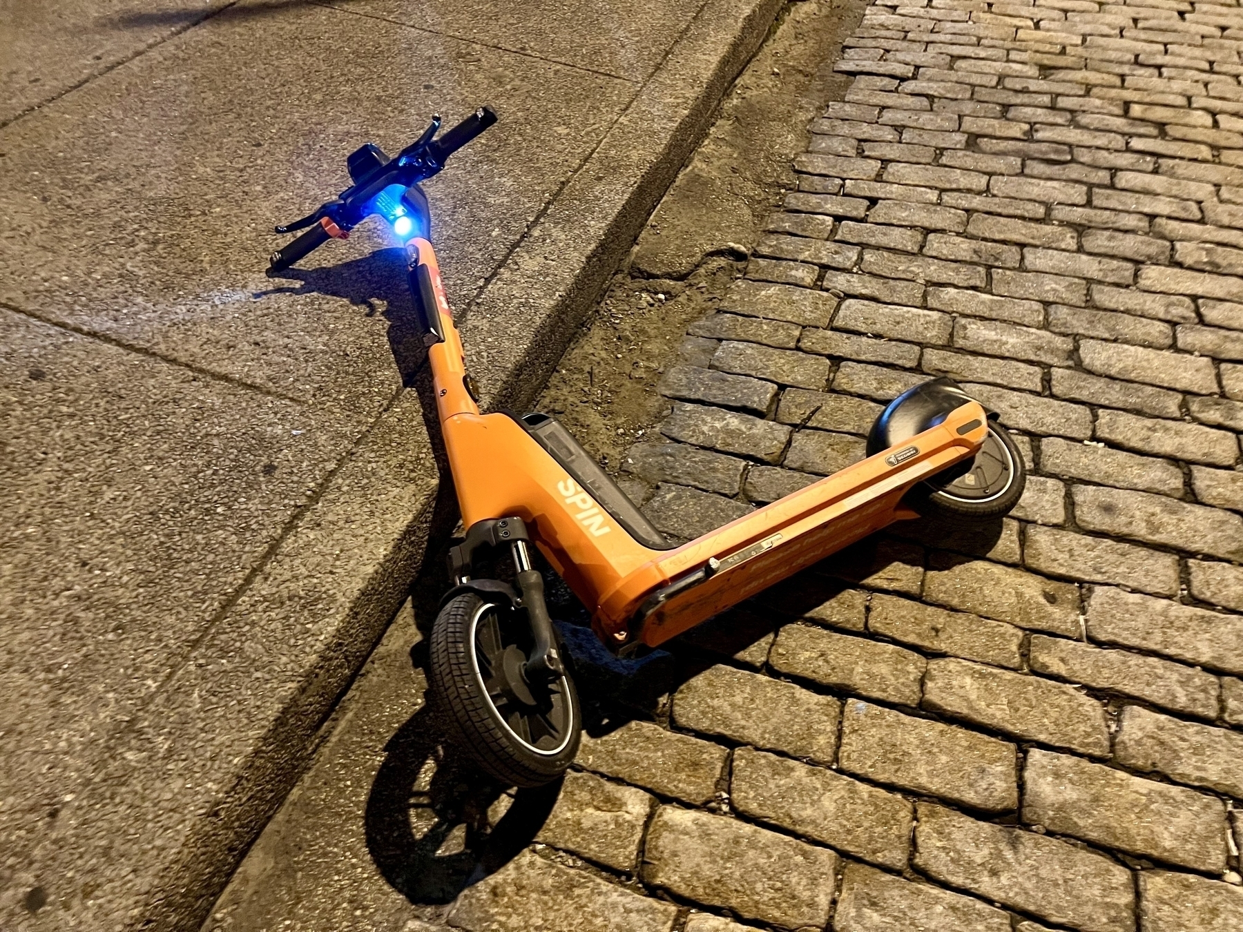 Broken electric scooter, lying on a brick road, with its headlight still on.