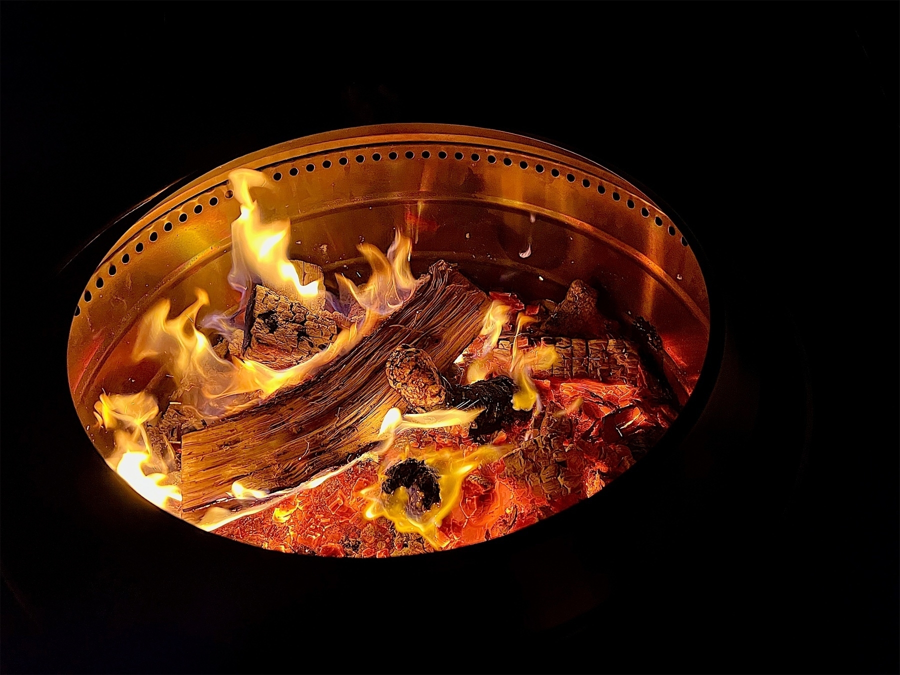 Fire contained within a metal fire-pit, with glowing embers and a few logs with flames licking upwards.