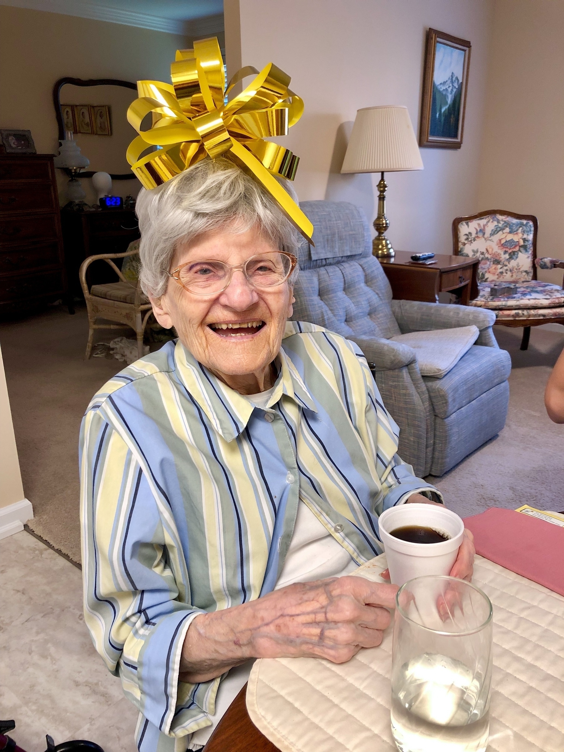 Little old Lady (don't worry, she'd have laughed at that joke) with glasses in a striped shirt (blue, yellow, and green), sitting in a chair at the kitchen table with a cup of coffee in her hand and a giant bow on her head, with a gigantic smile on her face.
