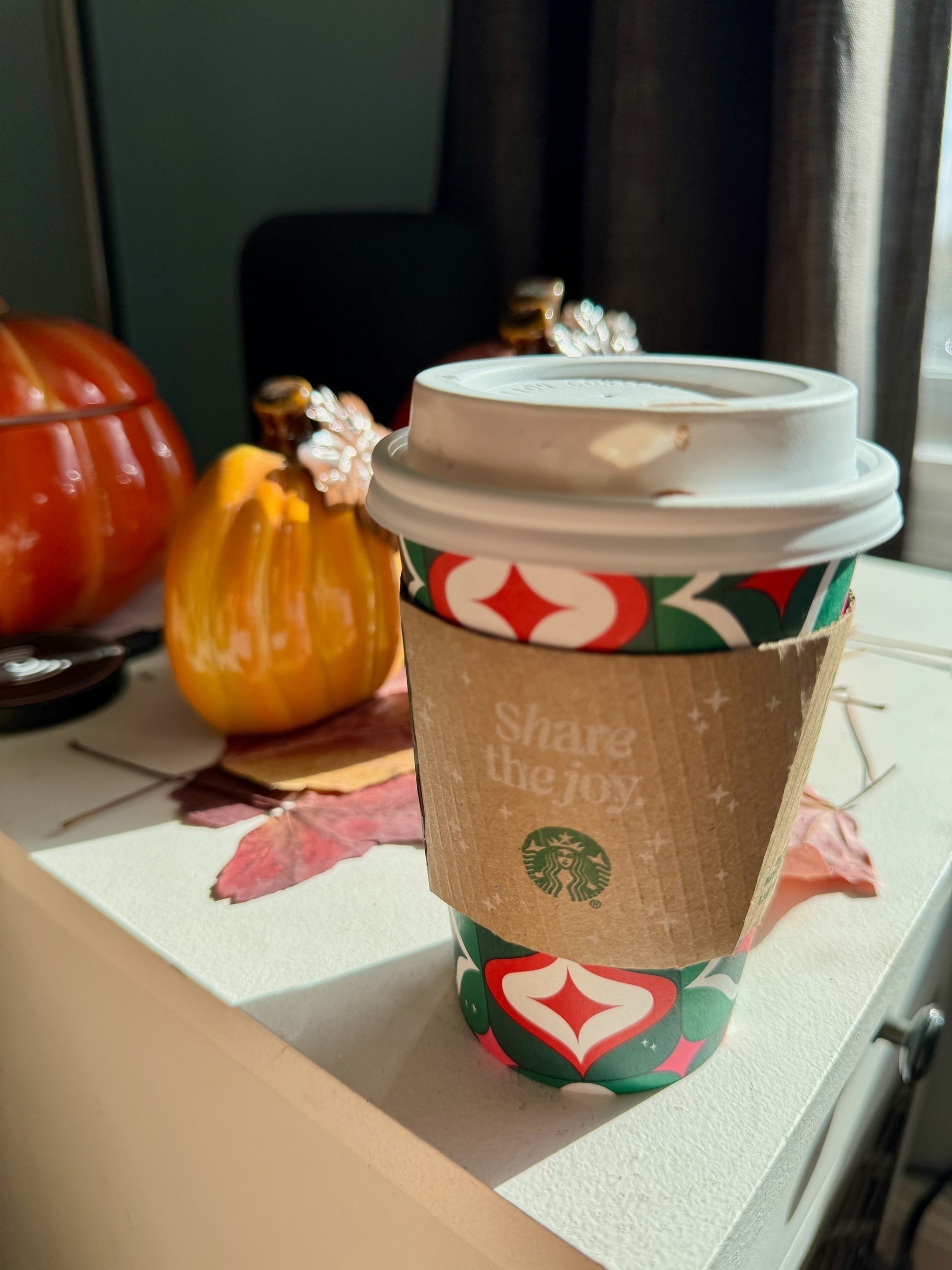 Paper coffee cup, with a Starbucks heat-sleeve, on a table, with ceramic pumpkins in the background and a wireless charging puck.