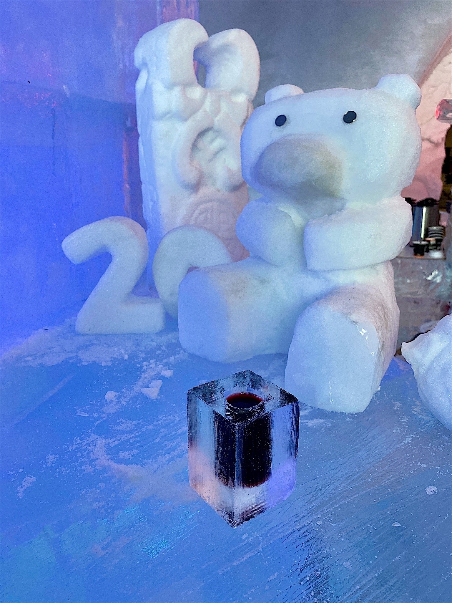 Rectangular shot-glass made out of ice, with red liquid inside, in front of a teddybear-like ice sculpture on a bar made of ice, sitting next to another ice sculpture (a random carving) and the '2' and '0' also made out of ice.