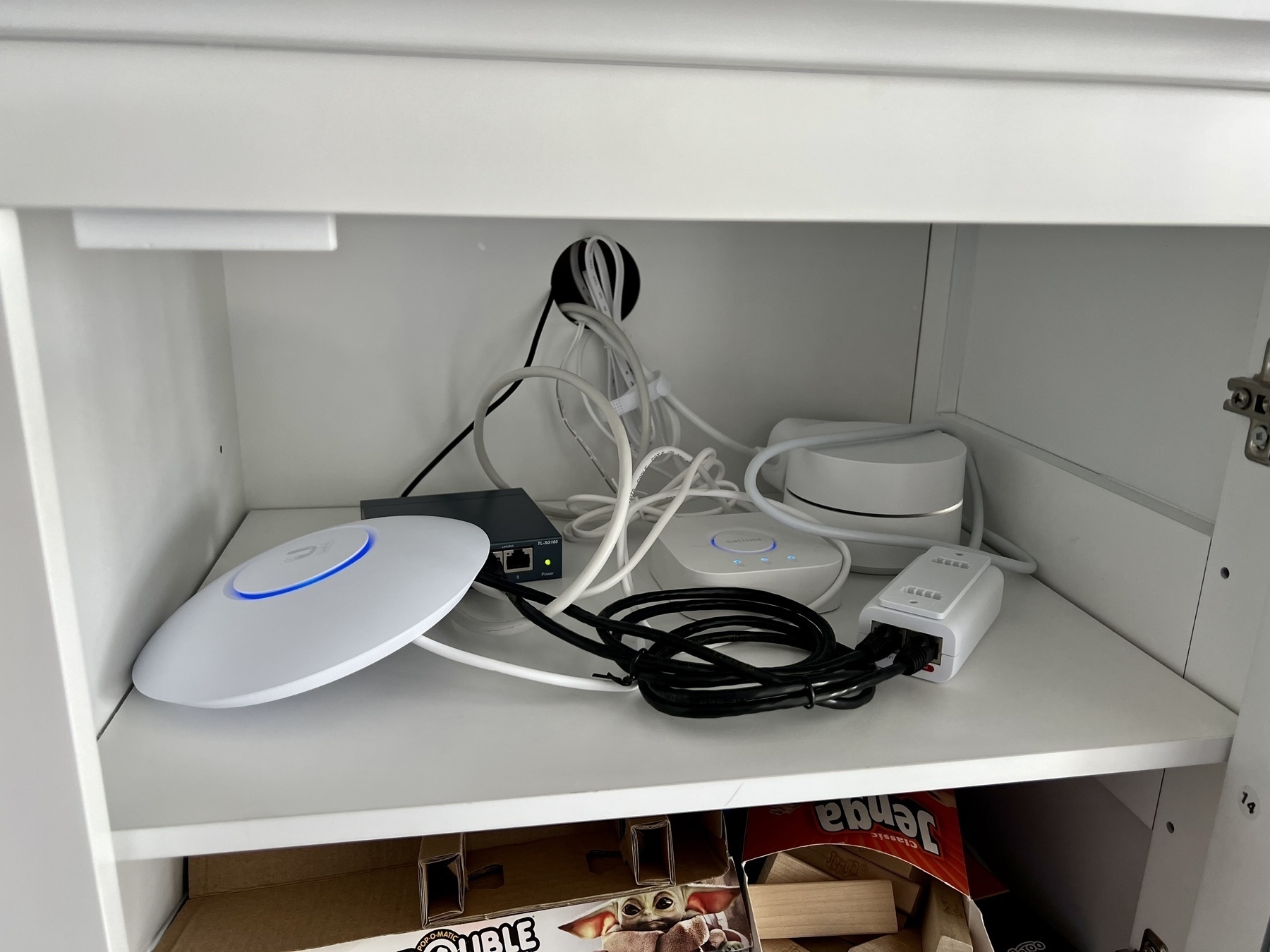 Lots of networking gear (Uni-Fi access point, networking switch, google access point, Phillips Hue hub, and PoE injector) on a shelf, inside a cabinet.