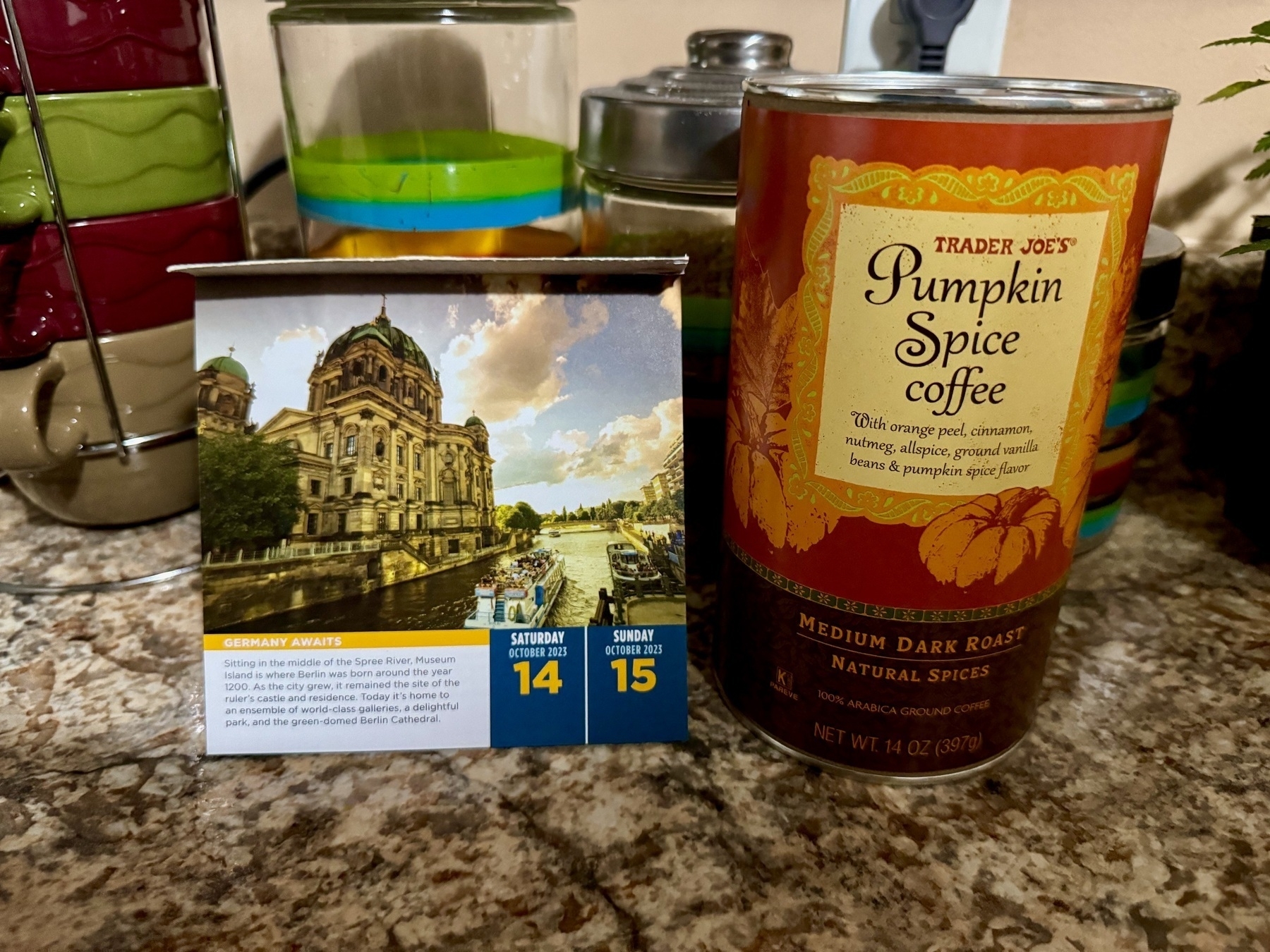 Photo of a container of Trader Joe's pumpkin spice coffee on the right side (on top of a kitchen counter, the 'fake' marbled pattern in various shades of brown), next to a calendar-book featuring a photo of a museum in Germany (it's a Rick Steve calendar).