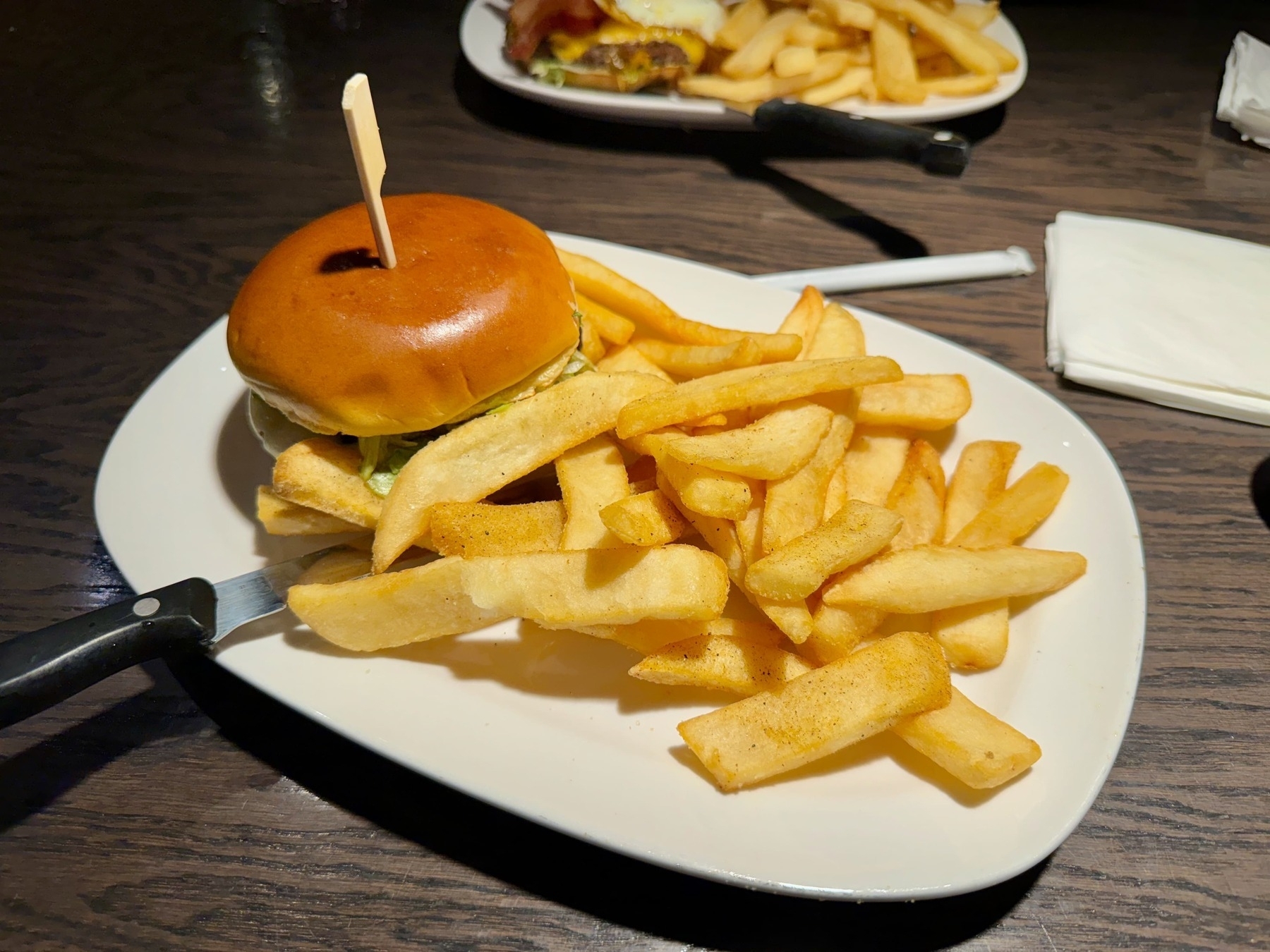 Two plates on a dark wood table, on two oval-ish white plates; fries on both of the plates but the plate in the foreground has a knife on it, as well as a burger.