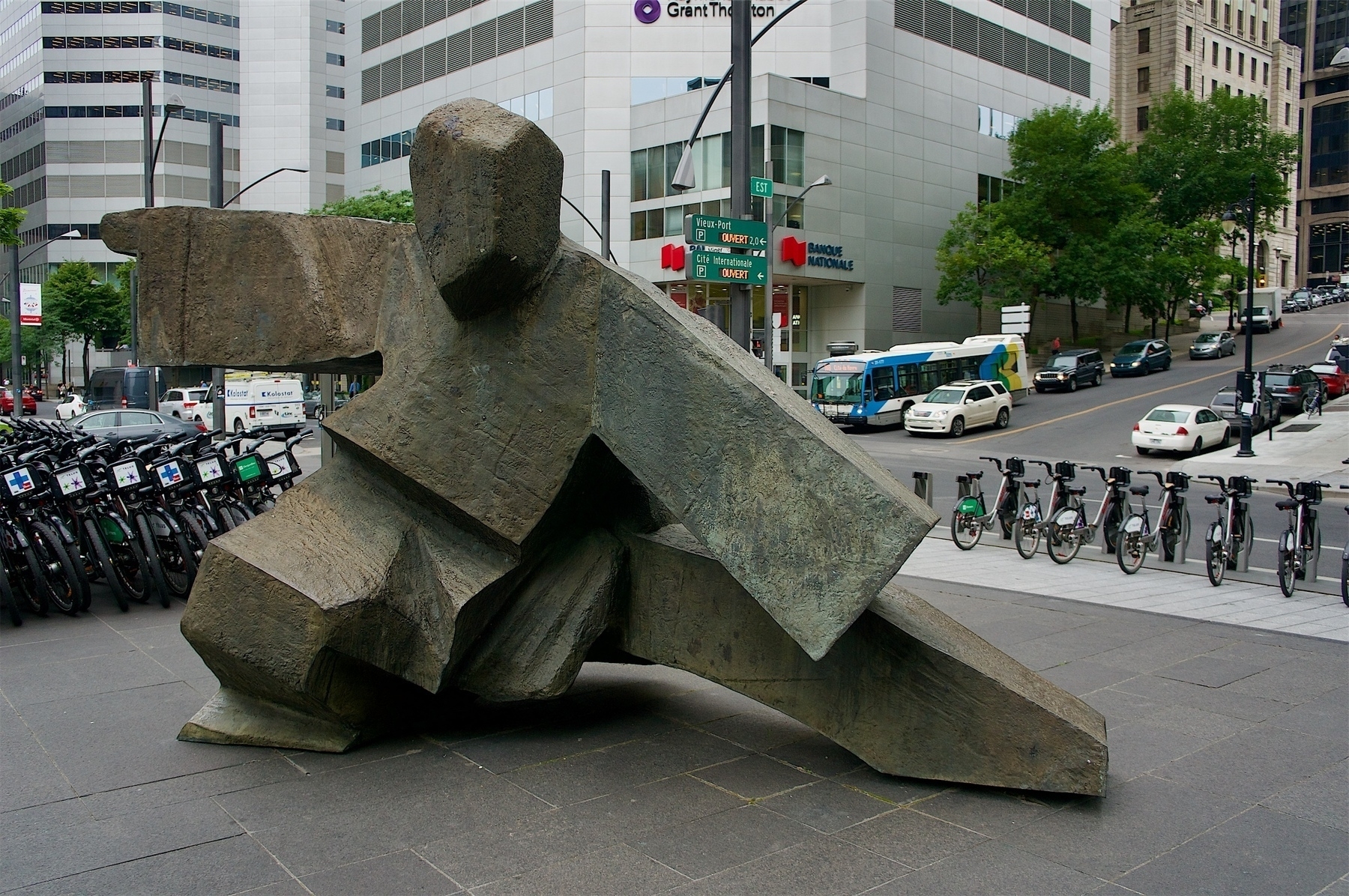 Stone statue in a tai chi pose, with public bikes in the background on the left and cars parked along a street going up a hill in the background on the right.