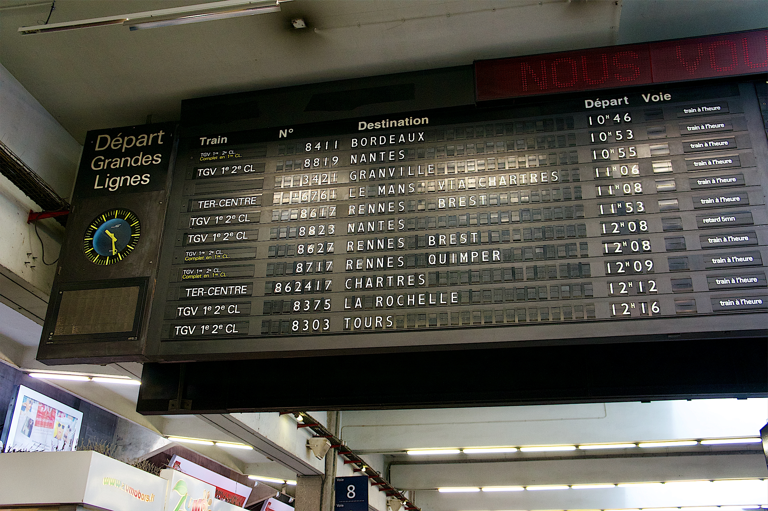 View of a split-flap display board (looking up from below) with times, places, and destinations in French, along with a clock on the left side of the board.