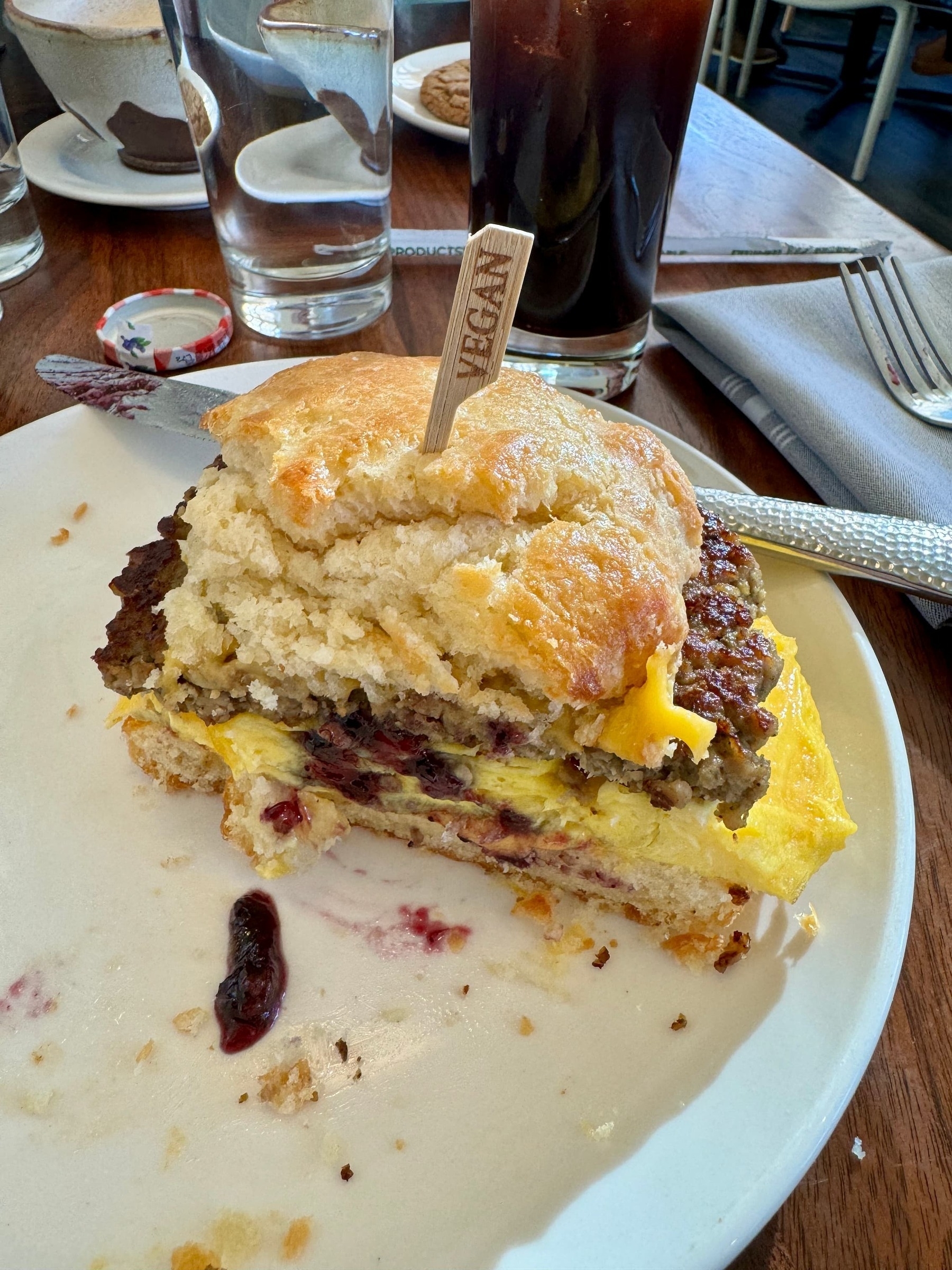 Breakfast sandwich (veggie burger, eggs, and cheese on a biscuit with a little bit of purple colored jam) with a wooden toothpick through the middle, the word 'Vegan' on it, on a plate. In the background is a glass of water and a glass of iced coffee.