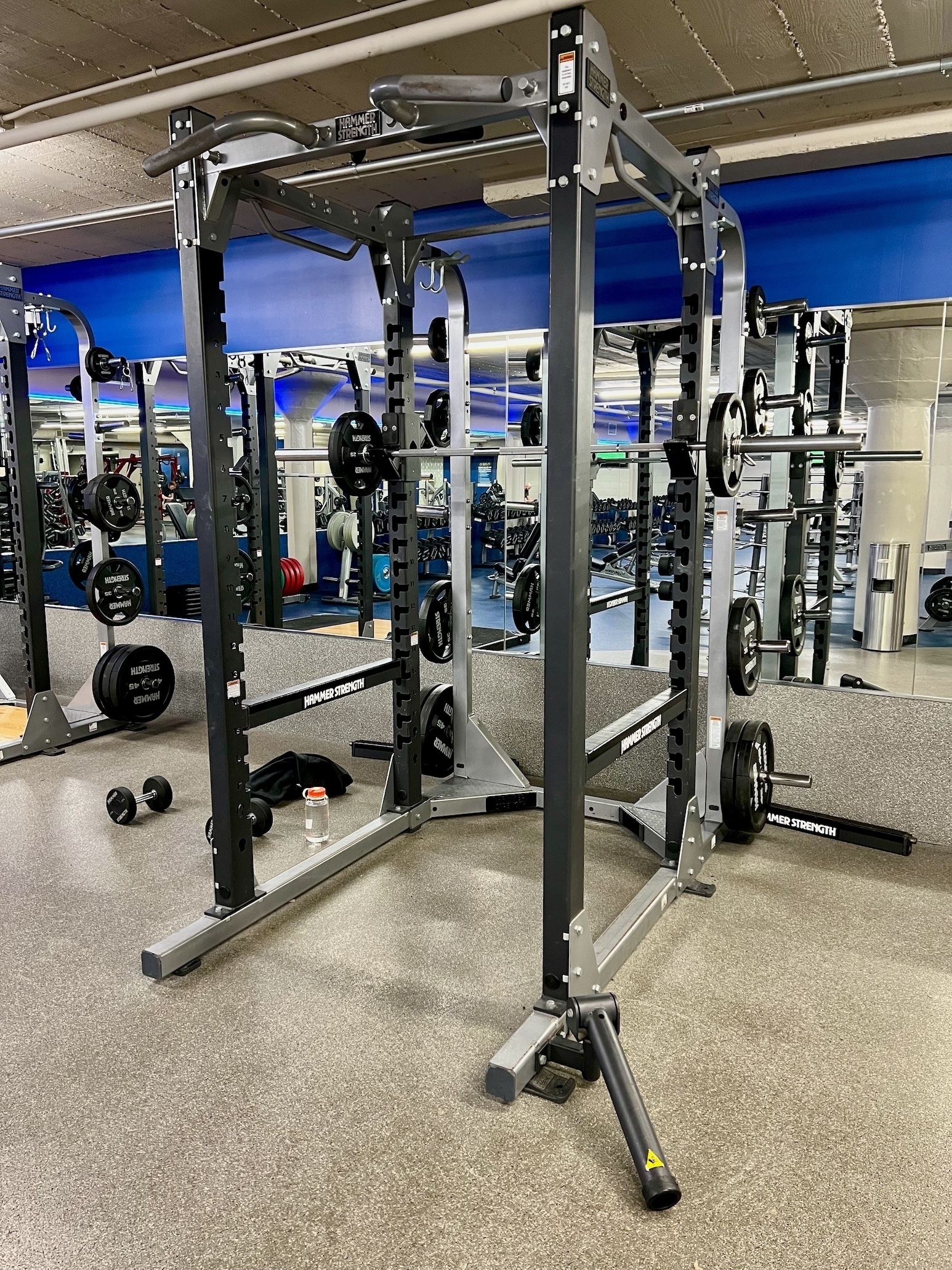 Weight machine, with various brackets to hold bar-bells, in front of a wall of mirrors, reflecting other parts of the gym; the machine is set up for squats.